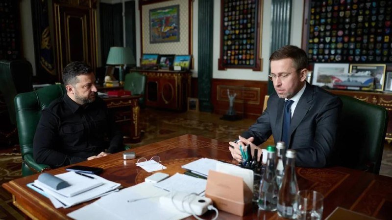Two men in business attire sit across from each other at a wooden desk in an ornate, decorated office, with papers, water bottles, and a power strip on the desk between them.