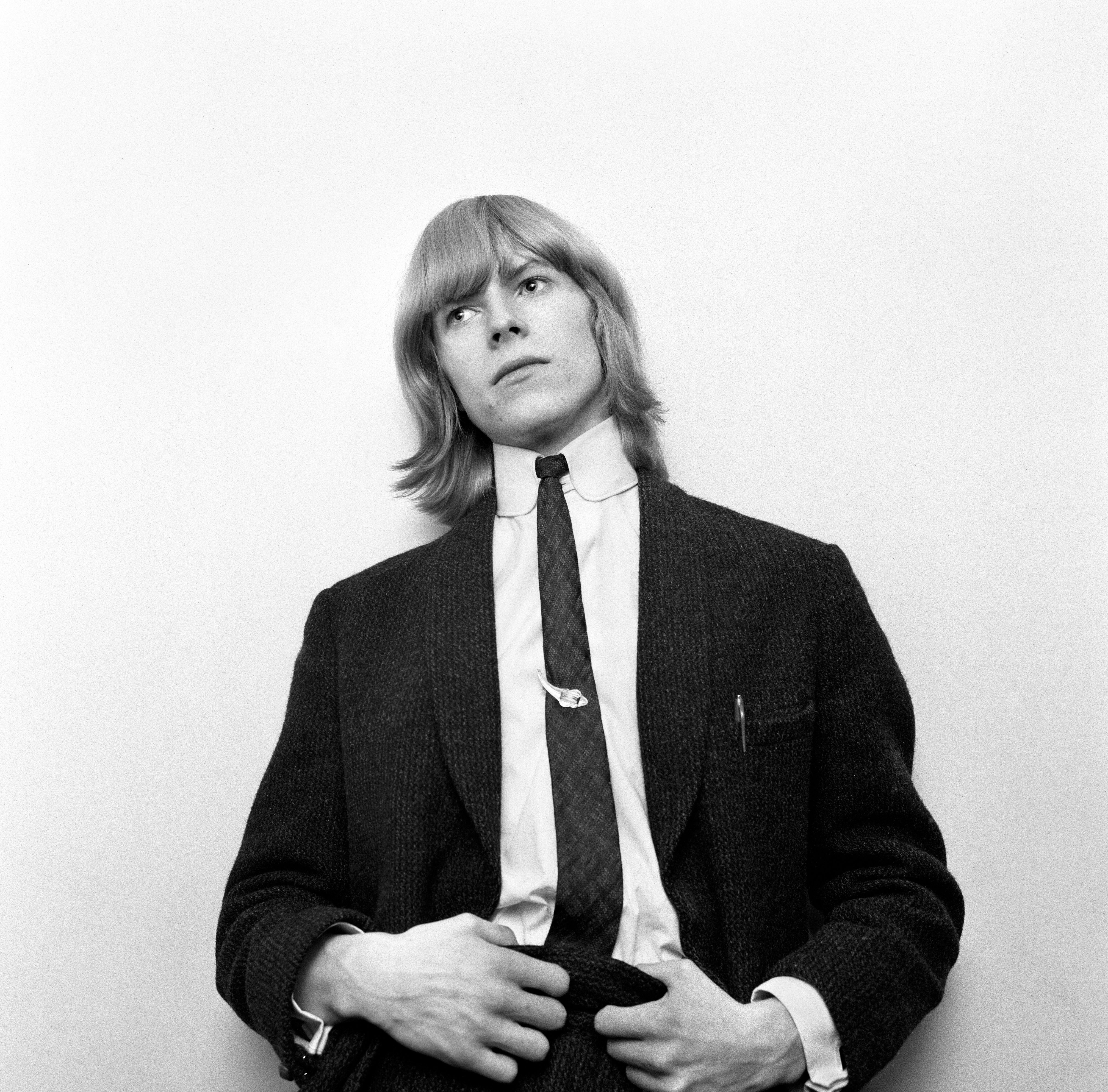 Black-and-white portrait of a young man with shoulder-length hair wearing a dark tweed jacket, white collared shirt, and dark tie, gazing to his right against a plain light background.