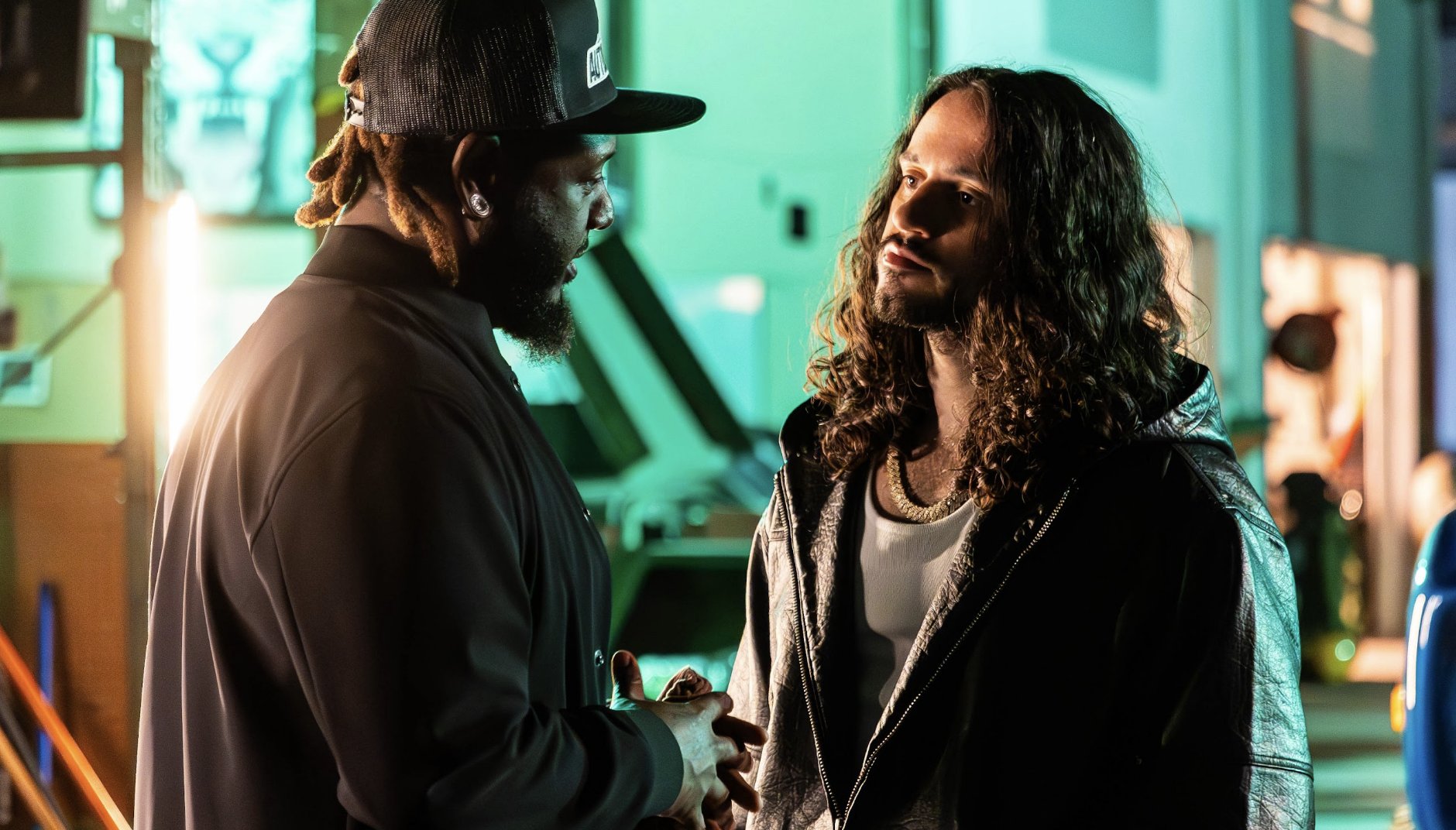 Two bearded men, one wearing a black cap and the other with long curly hair in a leather jacket, stand facing each other at night.