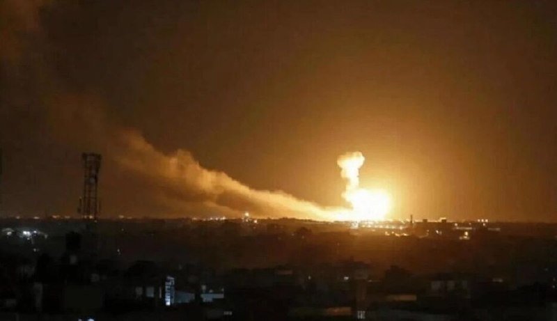 A bright orange fireball and thick smoke rise over a nighttime cityscape, with a communication tower visible on the left.