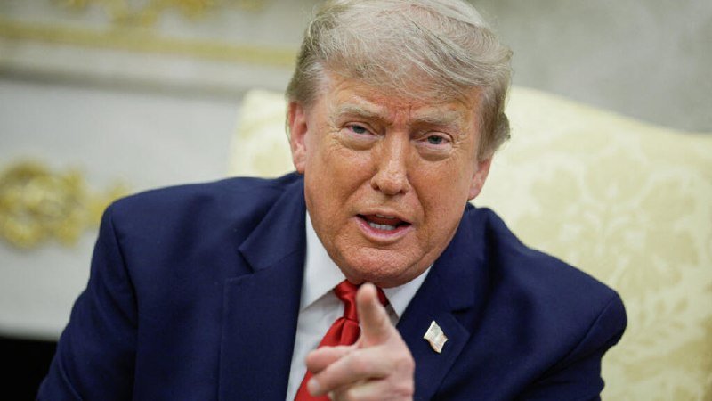 A man with blond hair wearing a navy suit and red tie gestures with his finger while seated in an ornate room with gold-trimmed furnishings.