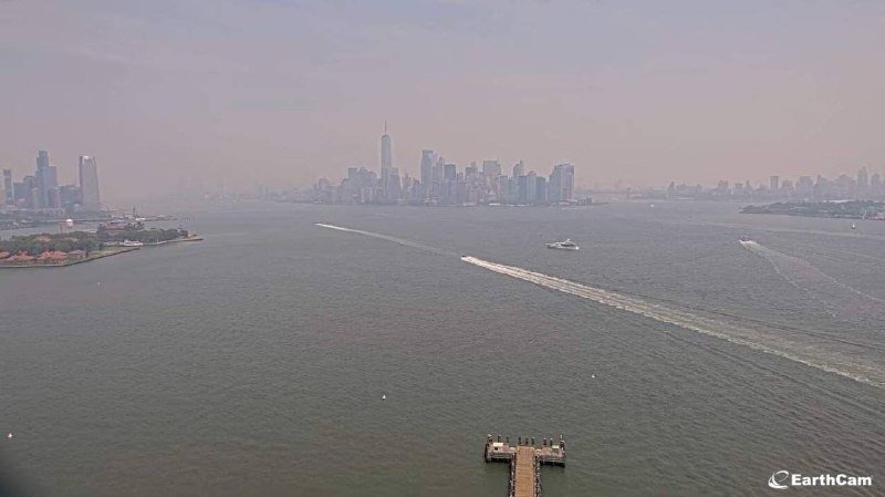 A hazy skyline of tall skyscrapers rises across a wide river, where a few boats leave white wakes near a small pier in the foreground.