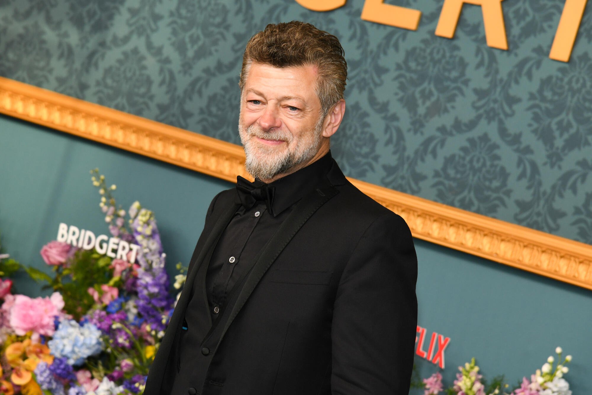 A gray-bearded man in a black tuxedo smiles at a "Bridgerton" premiere event, standing before a teal backdrop with floral arrangements.
