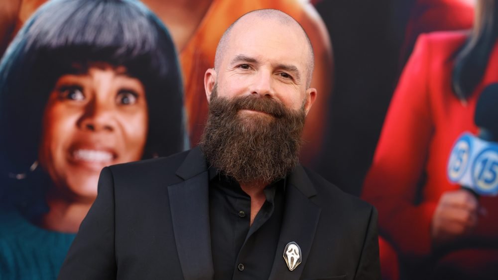 A bald, heavily bearded man in a black suit with a ghost-face pin smiles while standing in front of a movie poster backdrop.