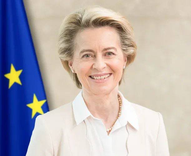 A smiling woman with short blonde hair in a light jacket stands beside a European Union flag.