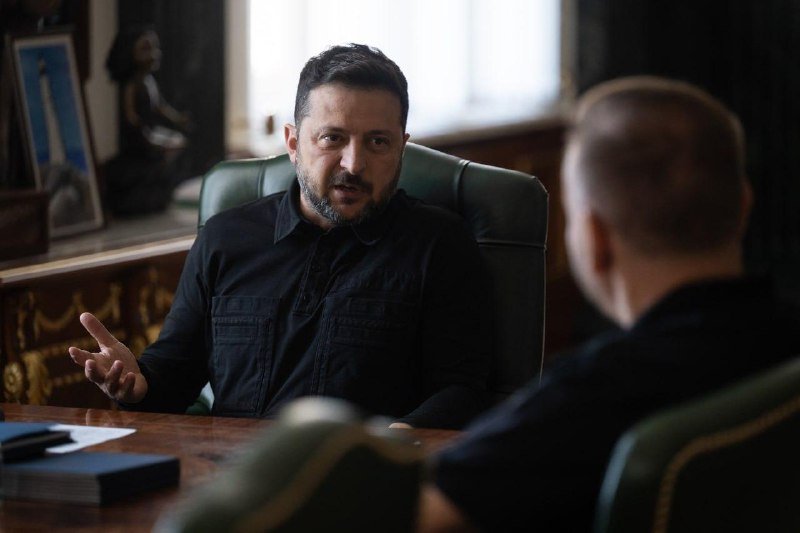 A bearded man in a dark shirt gestures with his hand while seated across from another person in a dark, ornately furnished room.