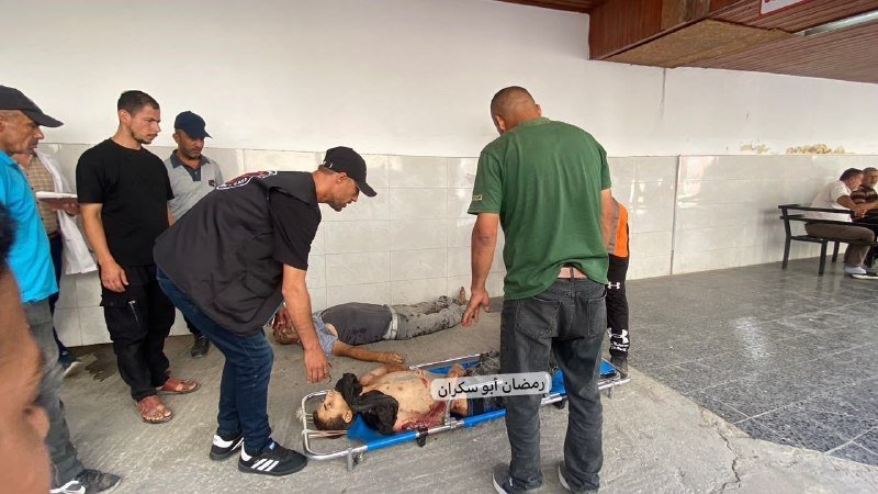 Several men gather around an injured person lying on a stretcher on a tiled floor inside a building, with visible injuries on the person's torso.