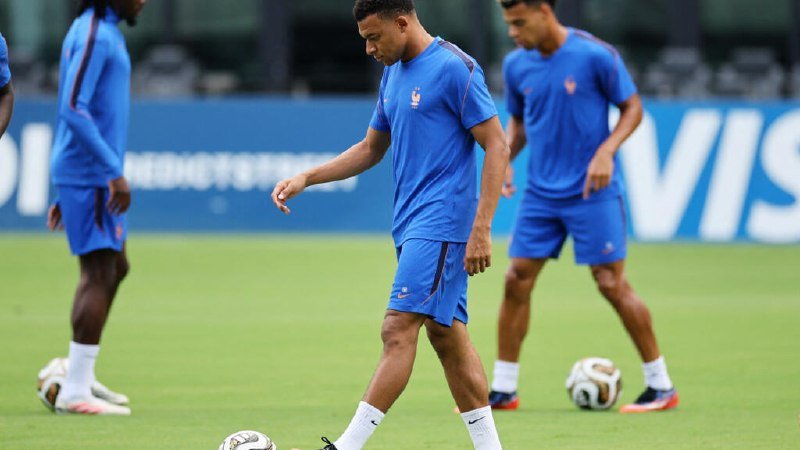 Three soccer players in blue uniforms with a rooster emblem walk across a grass pitch during a training session, with a "VIS" banner visible in the background.