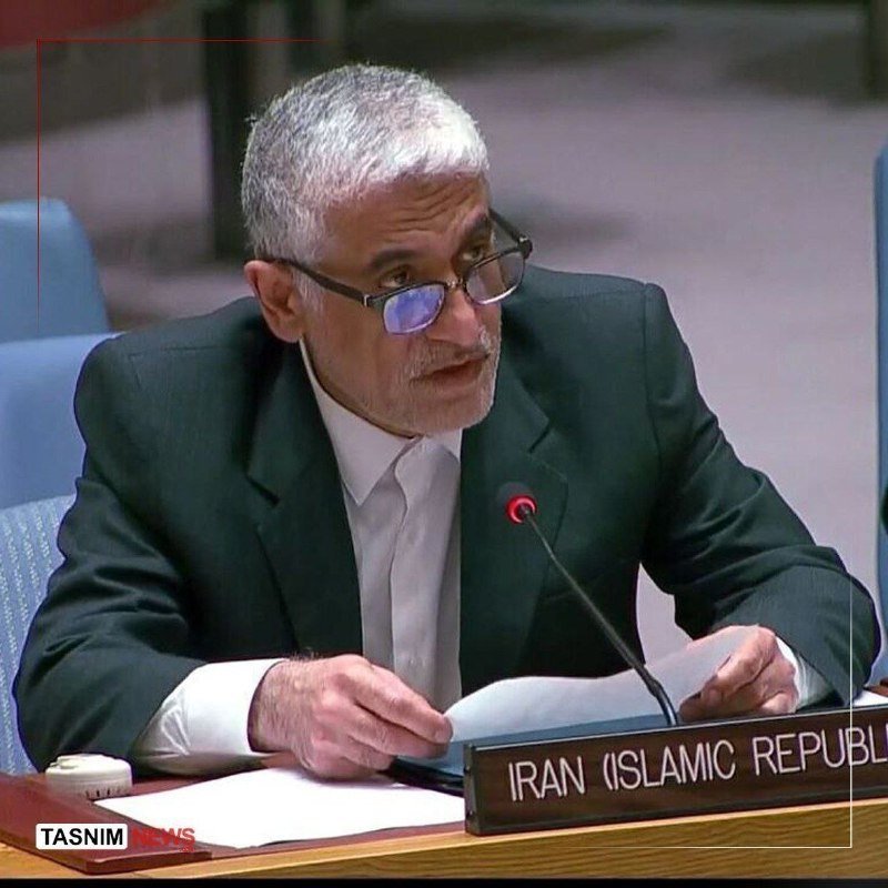 A gray-haired man in a dark suit and glasses speaks into a microphone at a desk labeled "IRAN (ISLAMIC REPUBLIC," holding documents.
