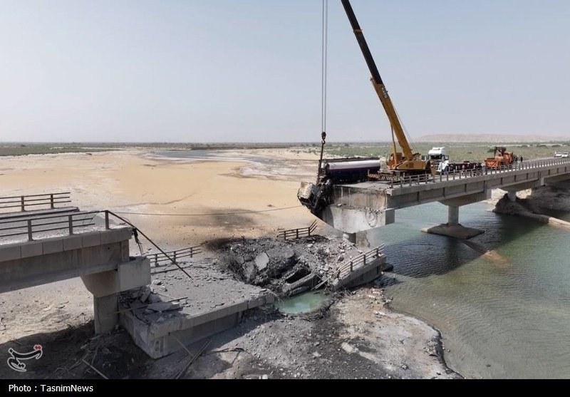 A crane on an intact bridge section dismantles a broken concrete bridge over a partially dry riverbed in a desert landscape.