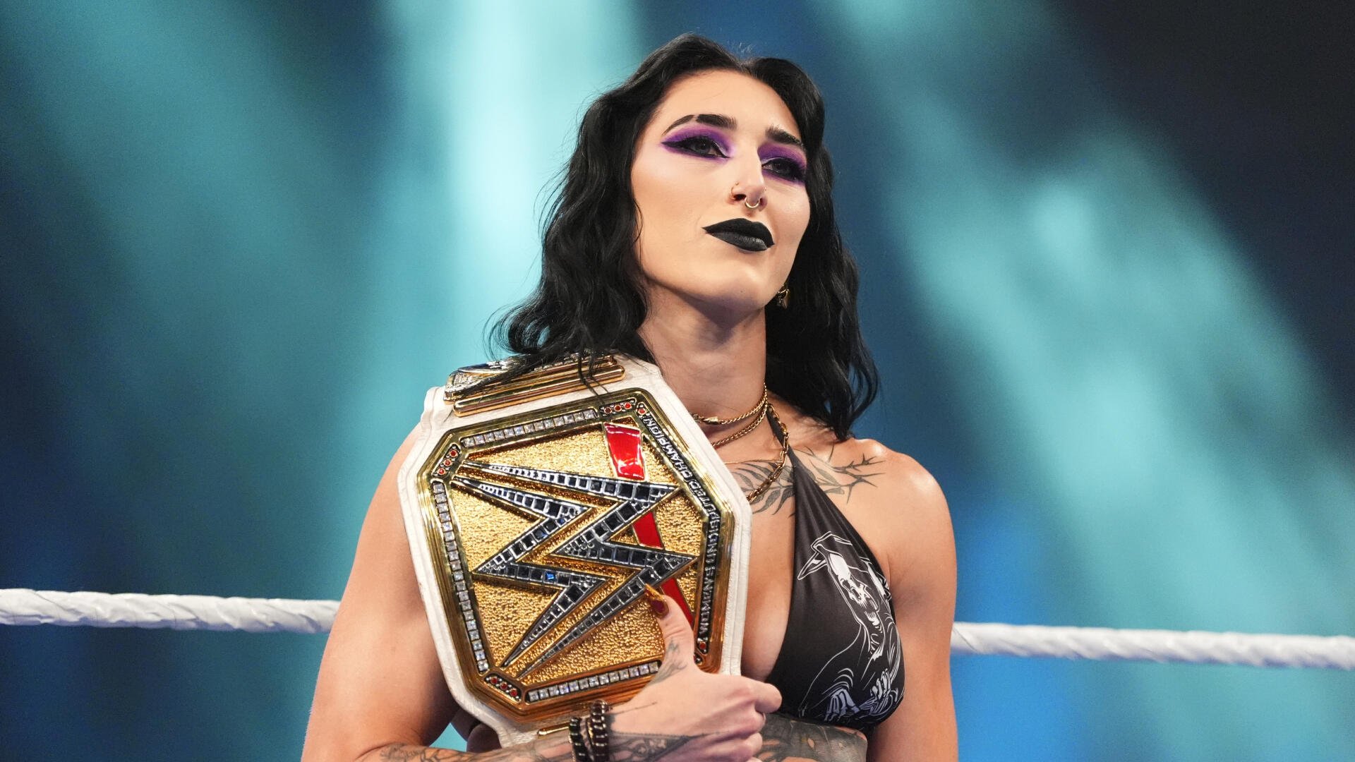 A woman with dark hair and purple eyeshadow stands in a wrestling ring holding a gold WWE championship belt over her shoulder.