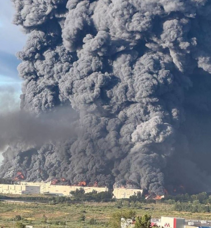 Massive plumes of dark gray smoke billow into the sky as bright orange flames engulf an industrial structure, with adjacent buildings, a grassy foreground, and a roadway also visible.