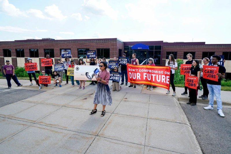 A woman speaks at a microphone while supporters behind her hold signs reading "Union Jobs," "Care Jobs," and "Don't Compromise On Our Future" outside a brick building.