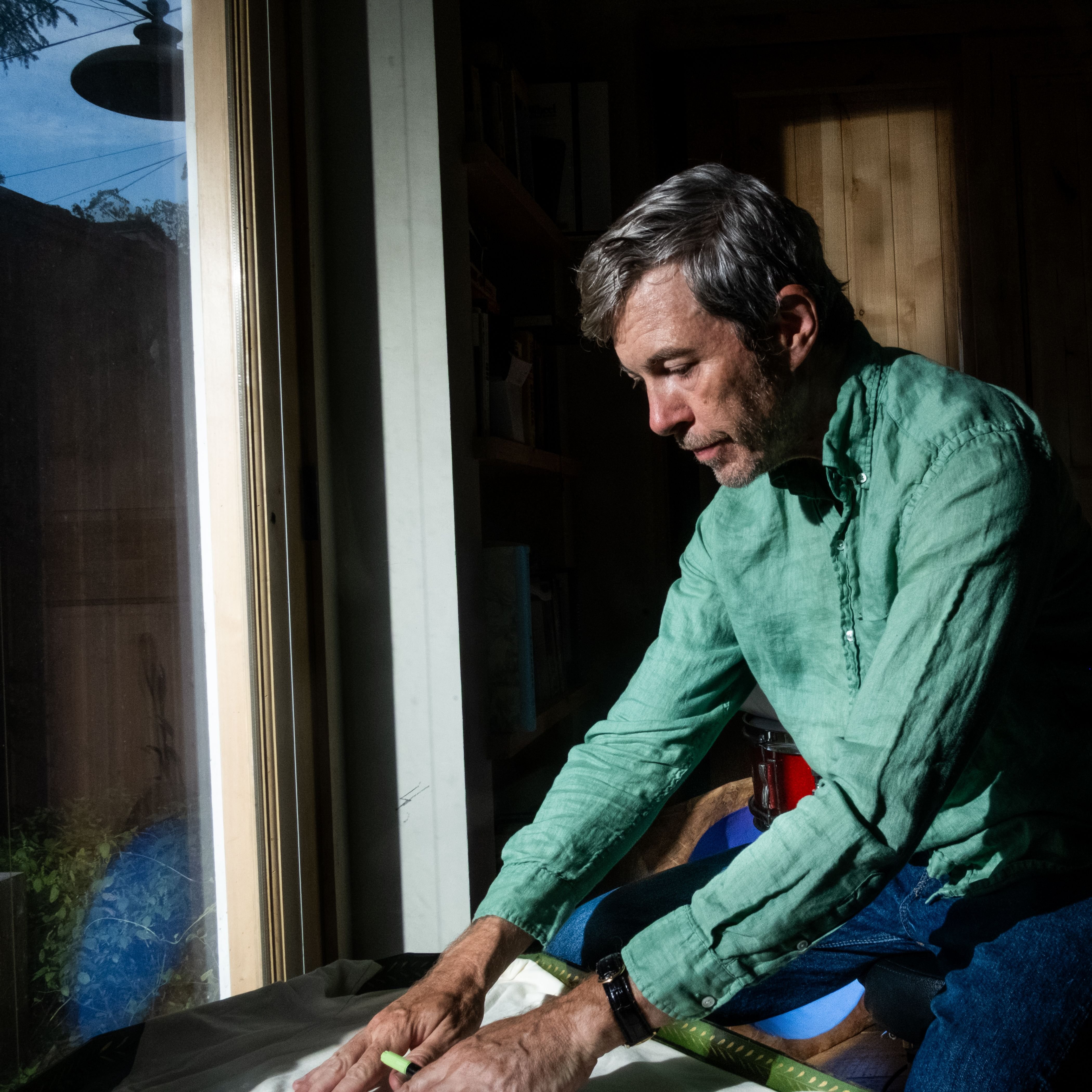 A gray-haired man in a green button-up shirt and jeans sits indoors near a window, looking down as he folds dark fabric on his lap.