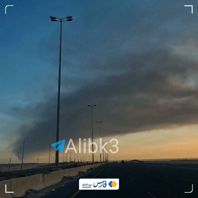 Dark smoke rises on the horizon beside a desert highway lined with tall streetlights under a dusk sky.