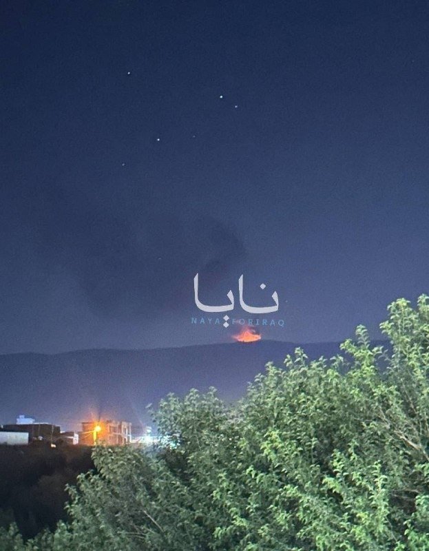 A nighttime view shows an orange fire burning on a distant hillside, with smoke rising into a dark sky above trees and illuminated buildings.