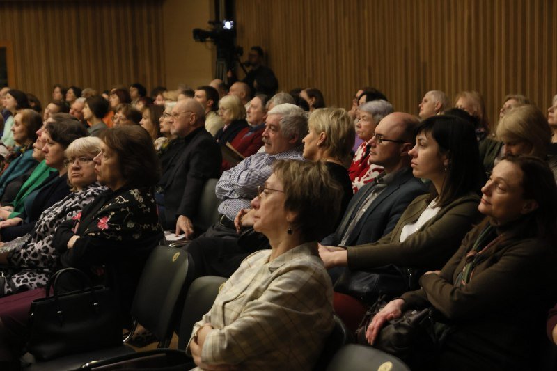 A seated audience attends an indoor event, with a video camera visible in the background.