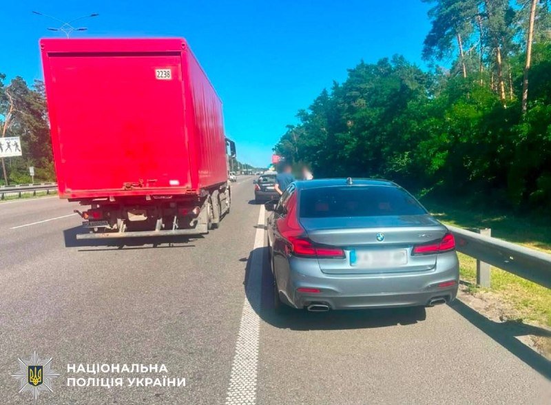 A red truck and a gray sedan are stopped on a sunlit highway next to a guardrail, with a police emblem and "Національна поліція України" text in the corner.