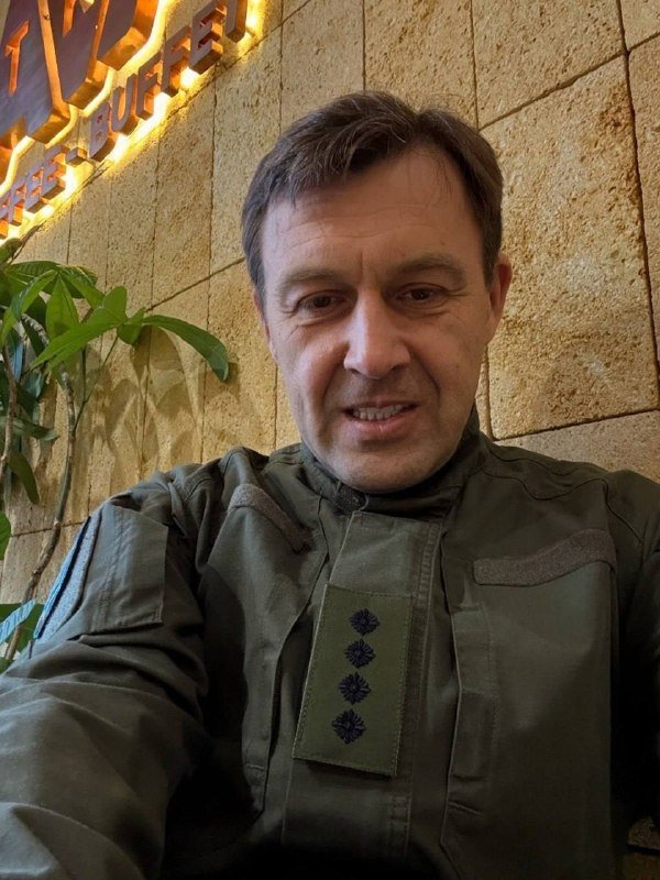 A man in a green jacket takes a selfie in front of a lit sign reading "FREE BUFFER" against a stone wall with greenery.