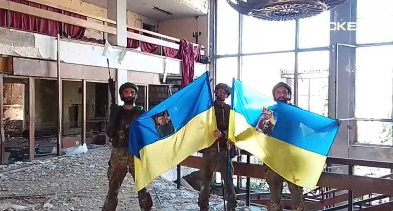 Three soldiers in camouflage uniforms and helmets stand inside a heavily damaged building, holding large blue and yellow Ukrainian flags and raising a fist in the air amid rubble and broken windows.