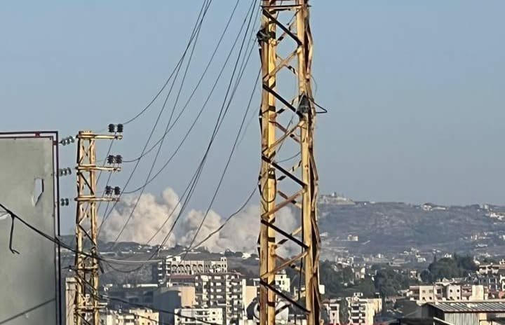 A yellow transmission tower stands in the foreground with power lines, while a plume of white smoke rises above a densely built hillside city.