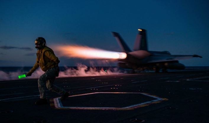 A crew member in flight gear runs across an aircraft carrier deck at dusk, holding a glowing wand, as a jet launches with afterburner flames and smoke.