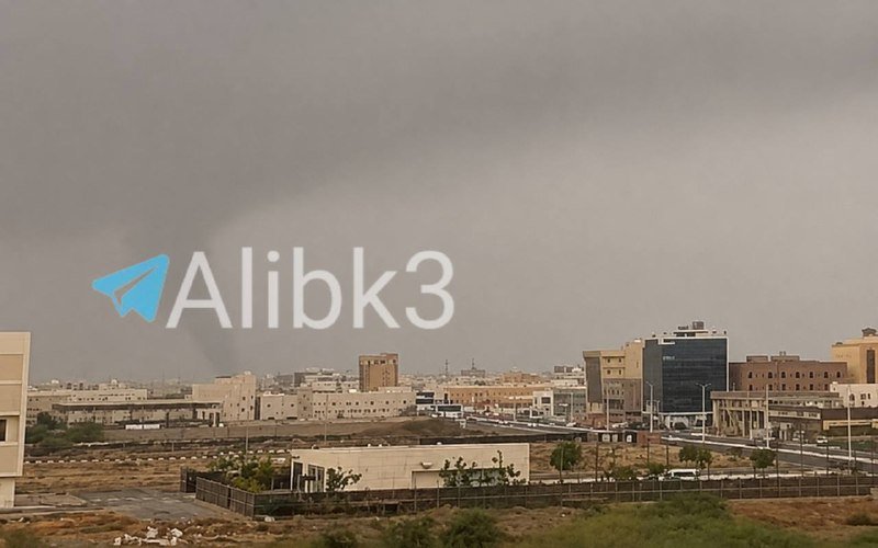 A dark funnel-shaped cloud descends from a stormy gray sky toward a cityscape of low-rise buildings under hazy conditions.