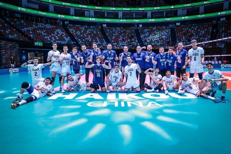 A volleyball team in white and blue uniforms poses together on a blue court marked "Haikou China" while holding a trophy inside an arena.