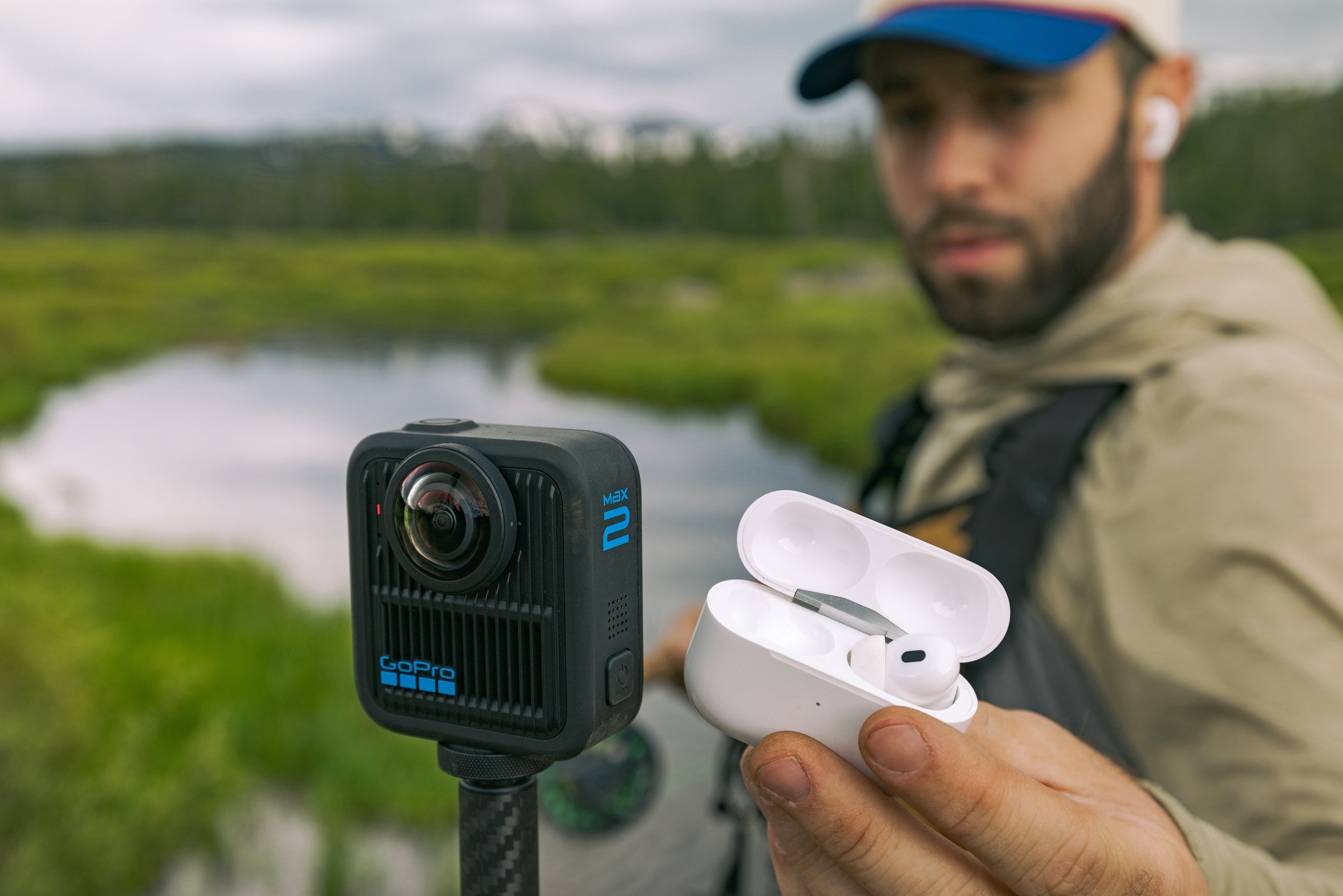 A man in a cap and beige jacket stands beside a marshy waterway, holding open a white earbud case next to a mounted GoPro Max camera.
