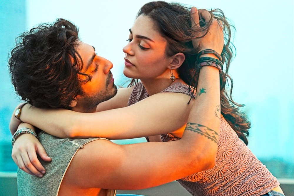 A man and woman embrace closely outdoors, their faces nearly touching, with visible tattoos on the woman's arm.