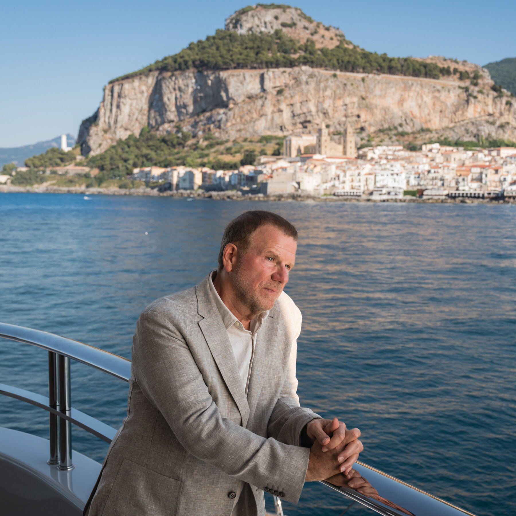 A man in a beige blazer leans against the railing of a boat, with a large rocky cliff and coastal town visible across the water in the background.