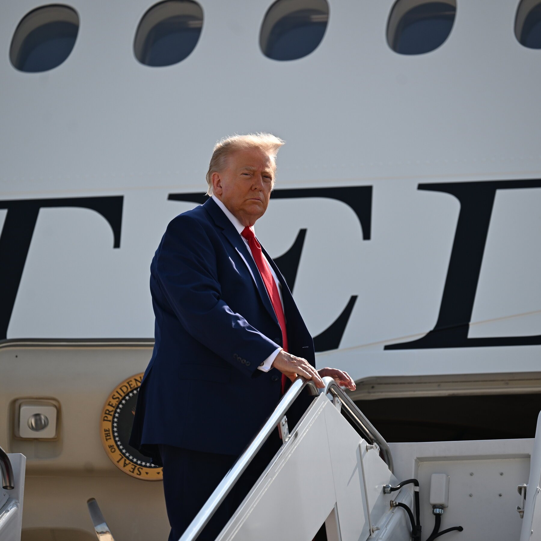 A man with blond hair, wearing a navy suit and red tie, descends the stairs of a white aircraft displaying the Presidential Seal and partial lettering.