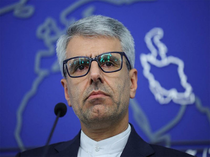 A gray-haired man wearing glasses and a dark suit stands before a microphone against a blue backdrop featuring a white map outline.