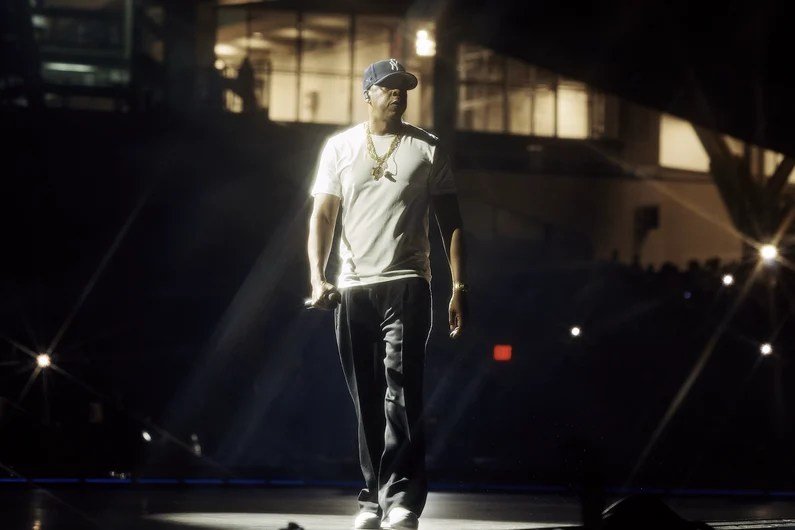 A performer wearing a white t-shirt, dark pants, gold chains, and a baseball cap walks across a spotlight-illuminated stage, holding a microphone.