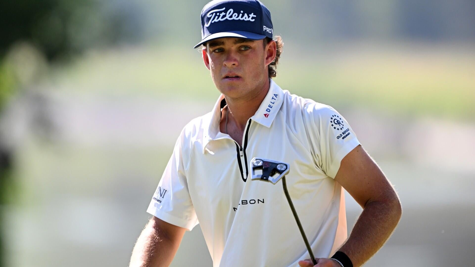 A golfer in a white polo shirt and Titleist cap holds a putter while standing on a sunlit course.