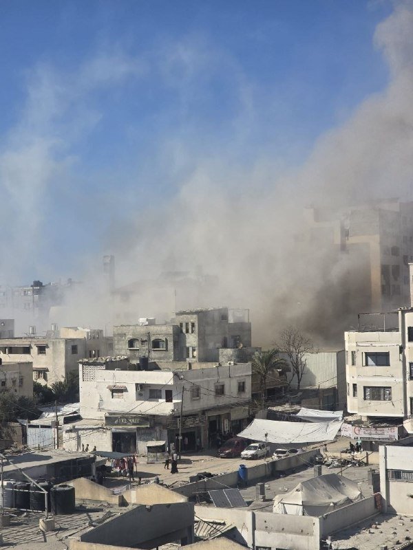 Plumes of gray smoke rise above densely packed multi-story buildings in a sunlit urban area, with people visible on the ground below.