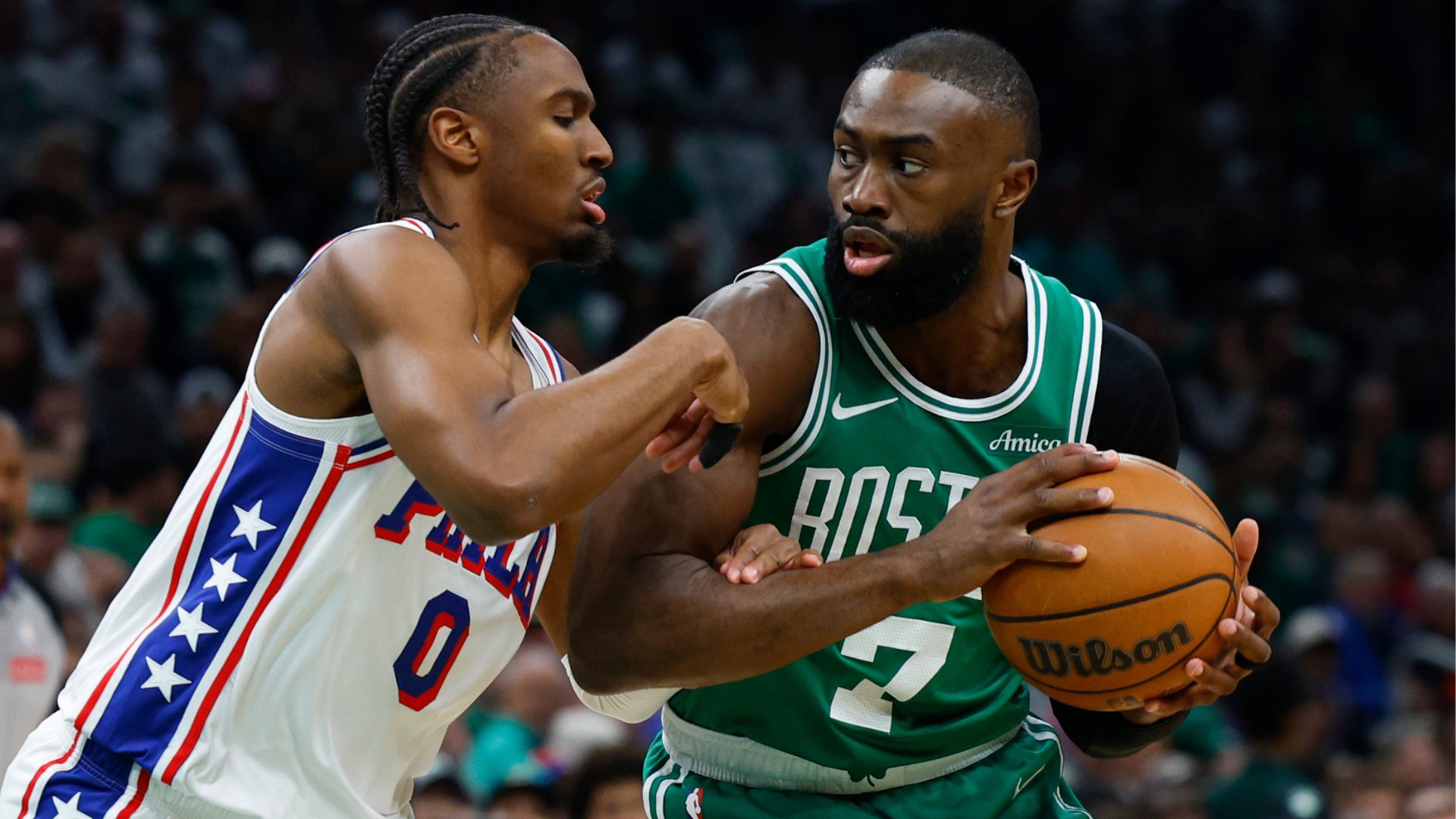 Two basketball players, one in a white jersey with "76ers" and stars and another in a green "Boston Celtics" jersey, compete closely for a Wilson basketball during a game.