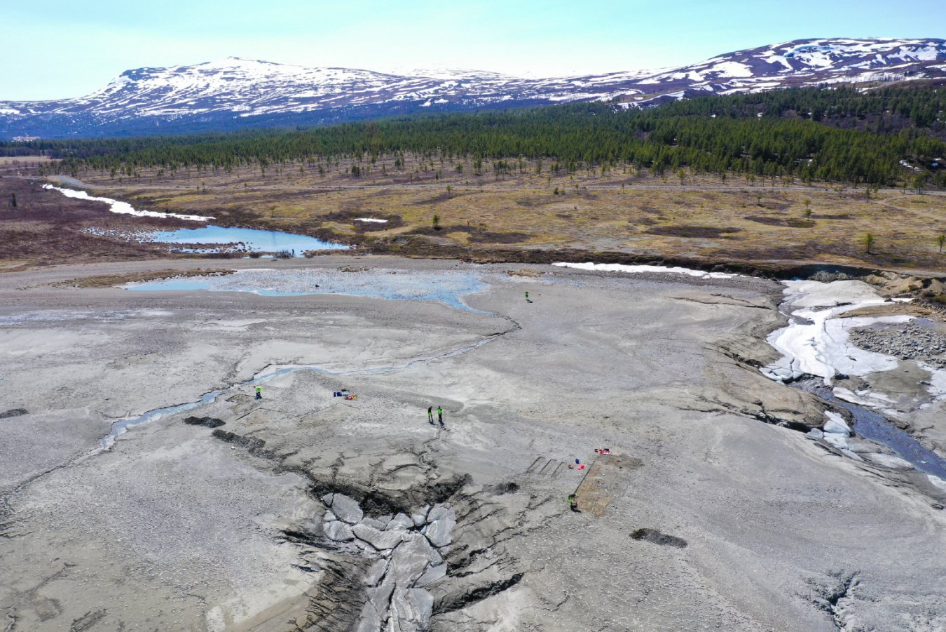 Aerial view of several people in high-visibility vests conducting fieldwork across a cracked, melting glacier with patches of snow and water, backed by forested terrain and snow-capped mountains.