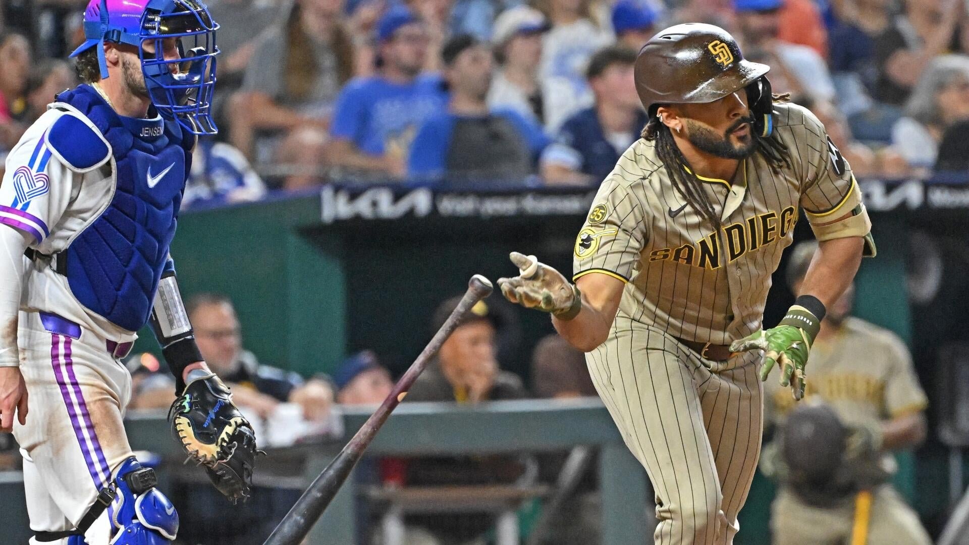 A San Diego Padres batter in a tan uniform reacts with open hands and a bat falling to the ground near a catcher in blue and white gear during a game.