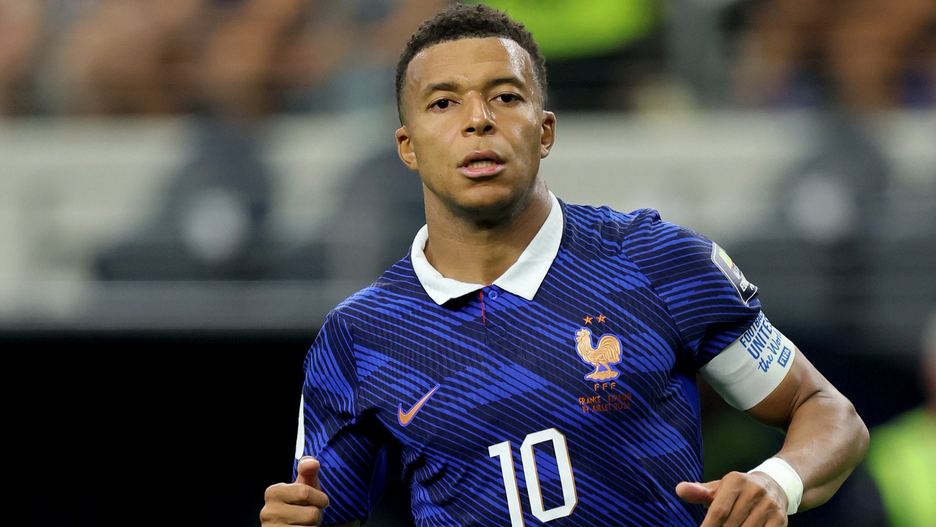 A young soccer player wearing a blue France #10 jersey and a United Against Racism captain's armband stands on a field with stadium seats blurred behind him.