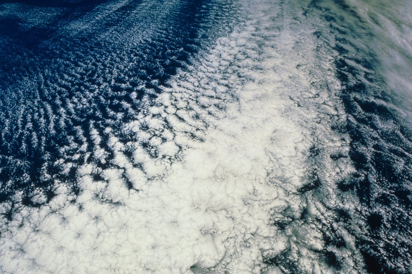 An aerial or satellite view shows a vast expanse of white cloud cover with a cellular, rippled pattern drifting over deep blue ocean waters.