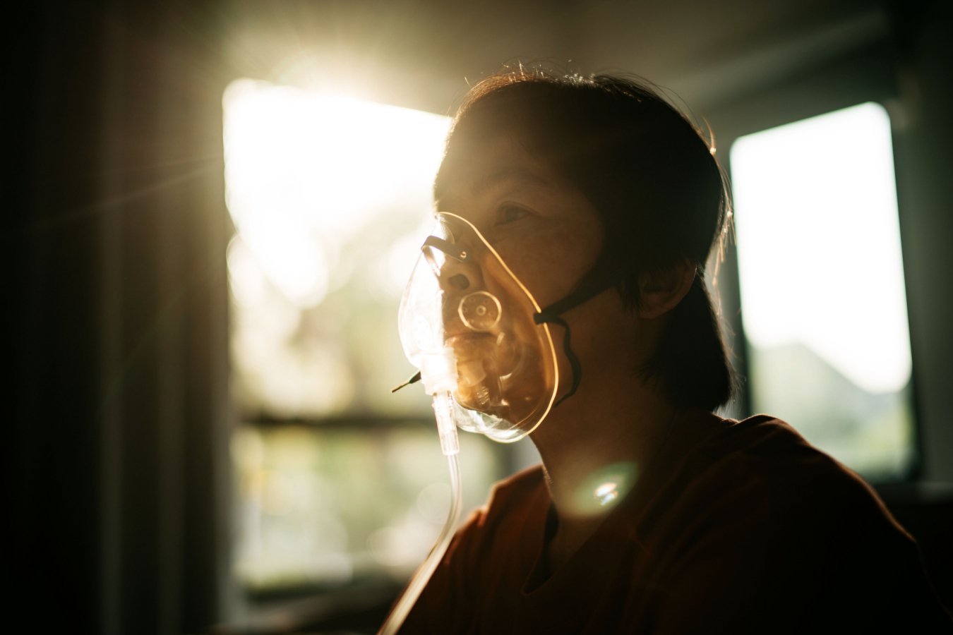 A person with short dark hair wears a clear oxygen mask, illuminated by bright sunlight streaming through a window behind them.