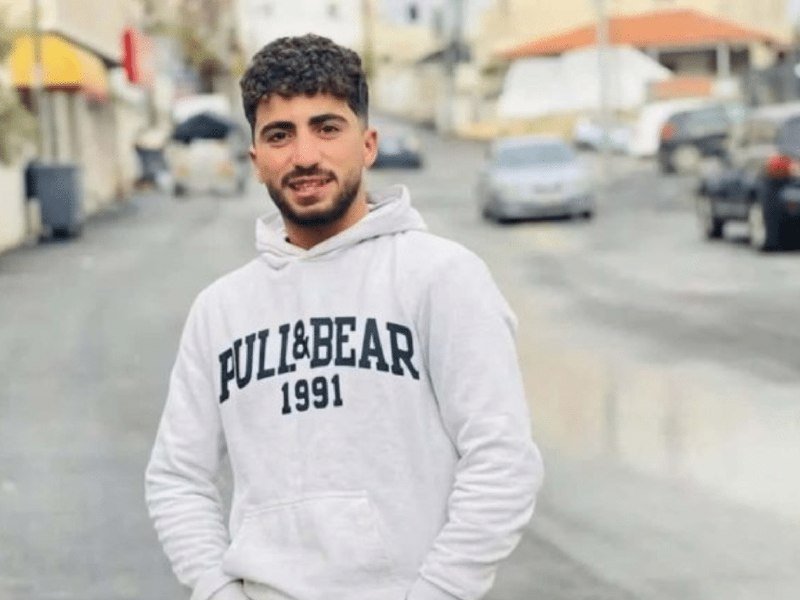 A young bearded man wearing a white "Pull&Bear 1991" hoodie stands smiling on a wet street with parked cars and buildings behind him.