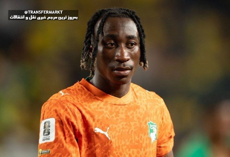 A male soccer player with dreadlocks wearing an orange Puma jersey featuring the Ivory Coast crest stands on a field with a blurred stadium crowd in the background.