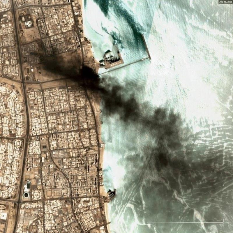 Satellite image dated July 15, 2020, shows a coastal urban area with dense black smoke billowing from a shoreline structure out over turquoise waters.