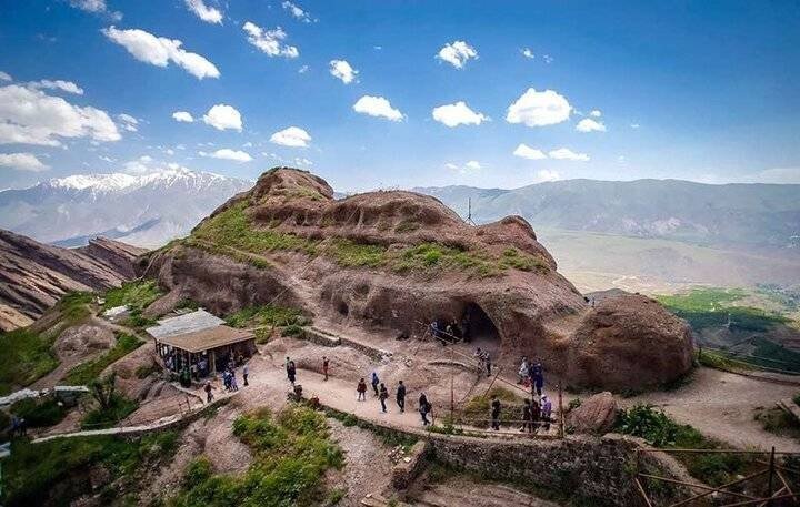 Visitors gather near a cave dwelling carved into a rocky mesa, with a small building and pathways overlooking a vast valley and distant snow-capped mountains beneath a partly cloudy sky.