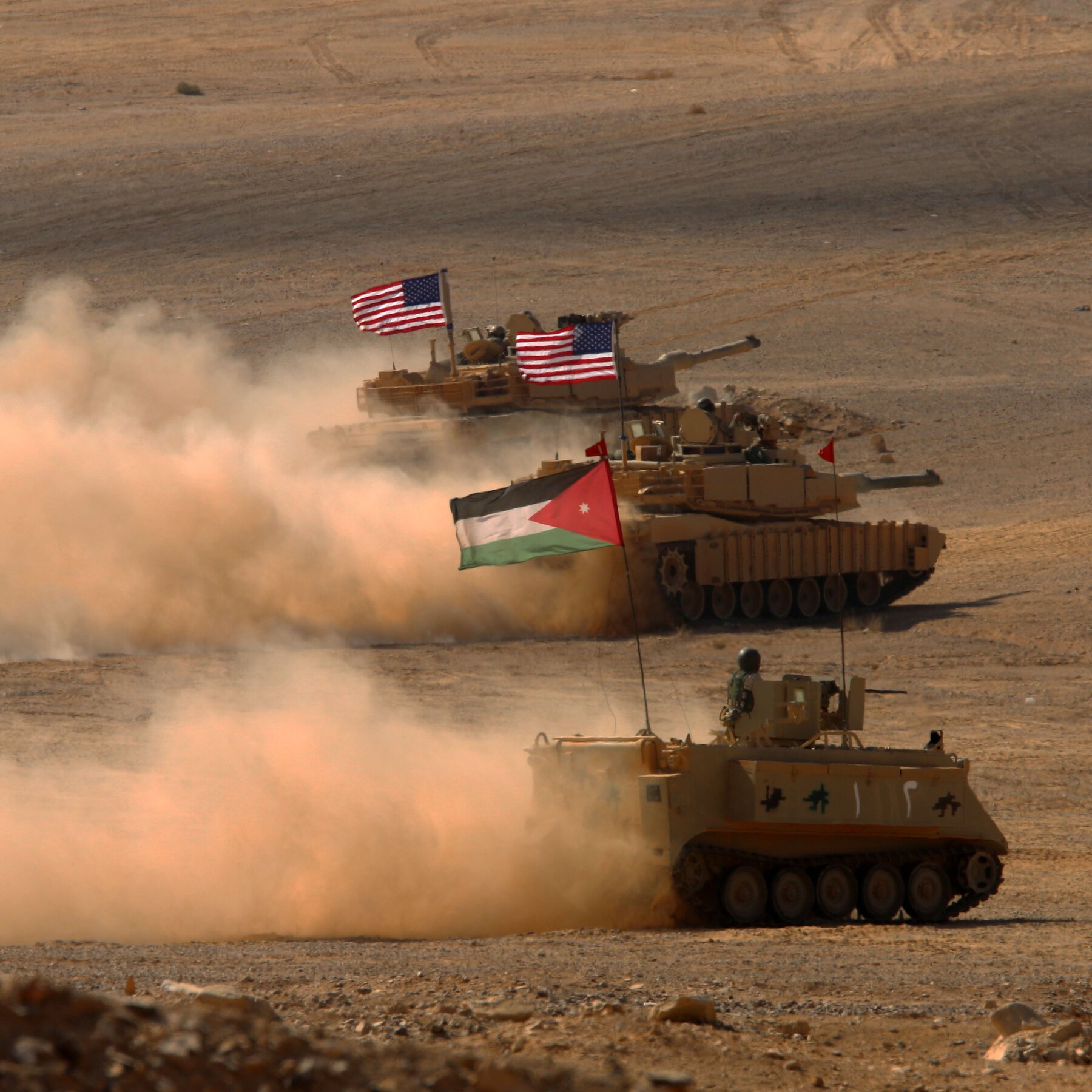 Tan military tanks and an armored personnel carrier kick up dust while advancing across a desert, flying American and Jordanian flags.