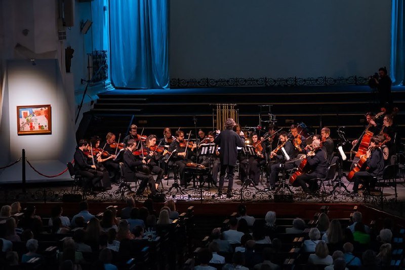 An orchestra in formal attire performs on a blue-curtained stage before a seated audience, conducted by a figure standing center-stage, with a framed abstract painting displayed nearby.