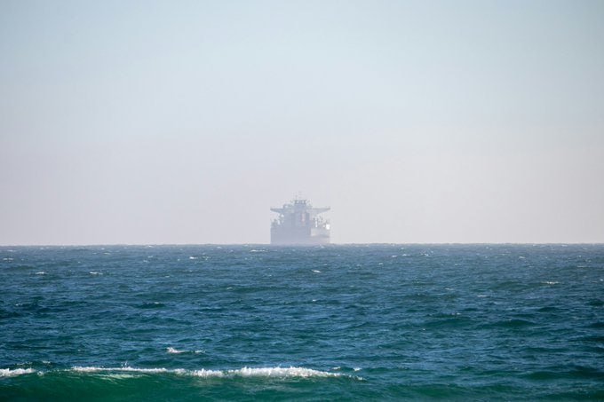 A large ship sits on the horizon, partially obscured by haze, in calm open ocean waters under a pale sky.