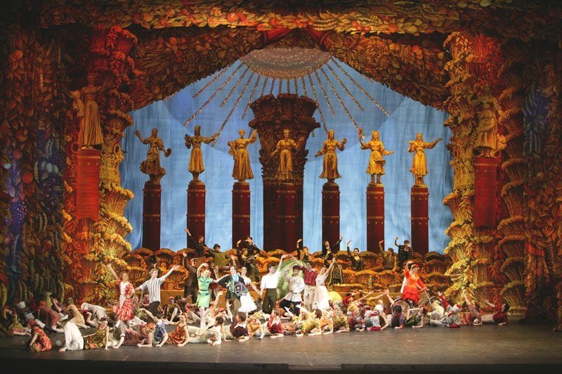A large ensemble of costumed performers dances on an ornate theater stage decorated with towering columns, golden statues, and elaborate autumn-themed set pieces.